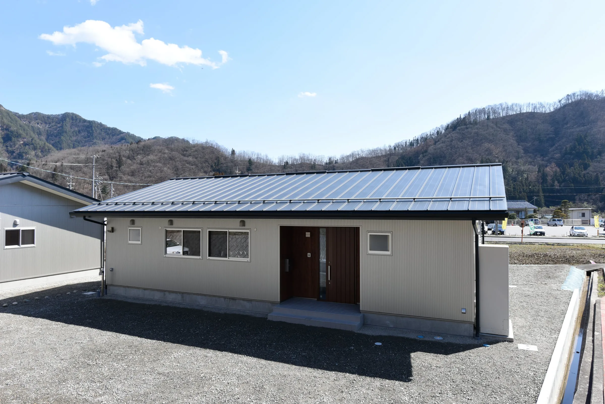 山を背にした平屋住宅の正面外観。金属屋根と木目の玄関扉