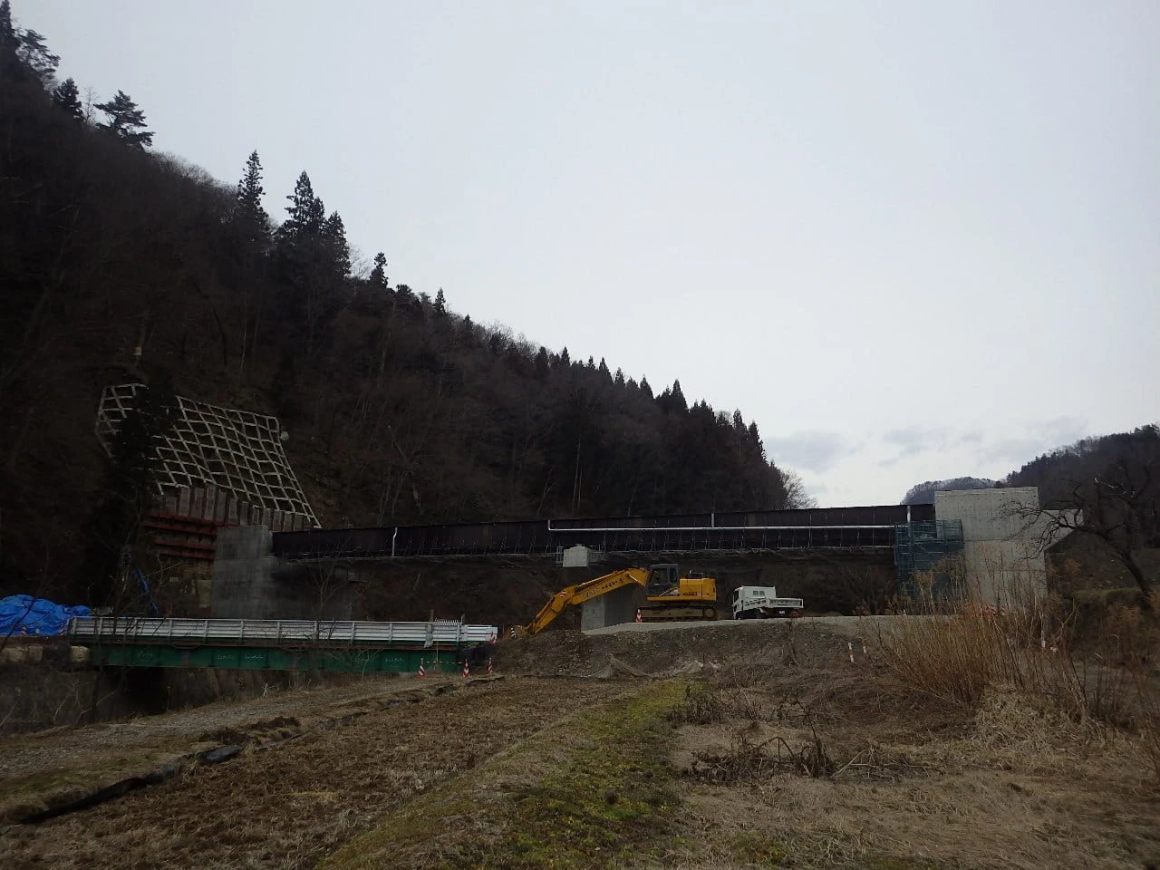 谷を渡る鋼桁と、たもとで作業する重機を対岸から見たところ