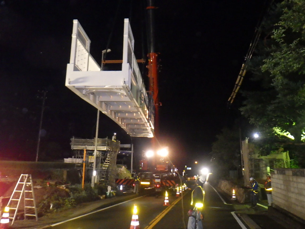 夜間、クレーンで吊り上げられた白い歩道橋の桁と、交通誘導中の道路