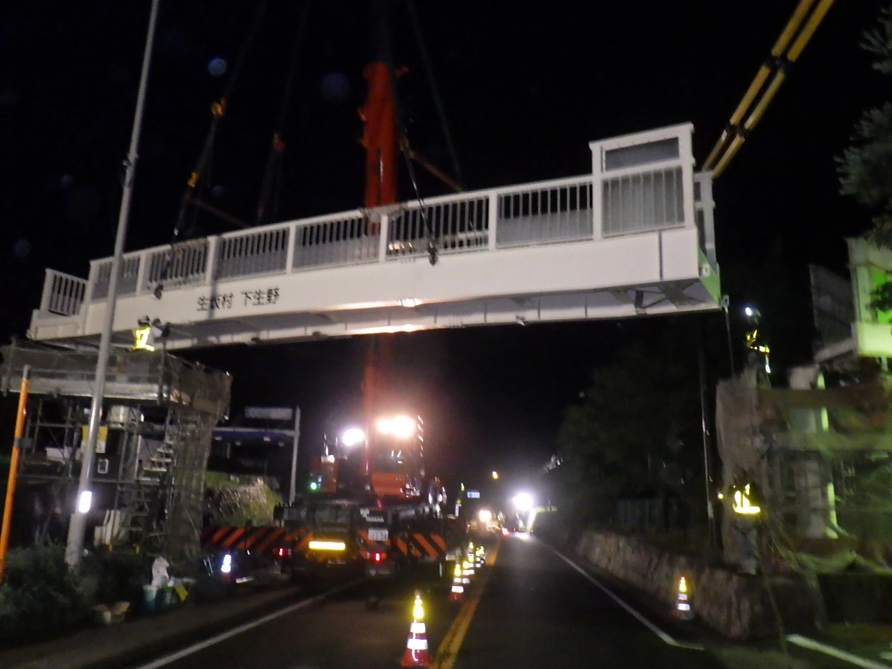 夜間、道路をまたぐ位置まで吊り上げた歩道橋の桁。側面に「生坂村　下生野」の銘板