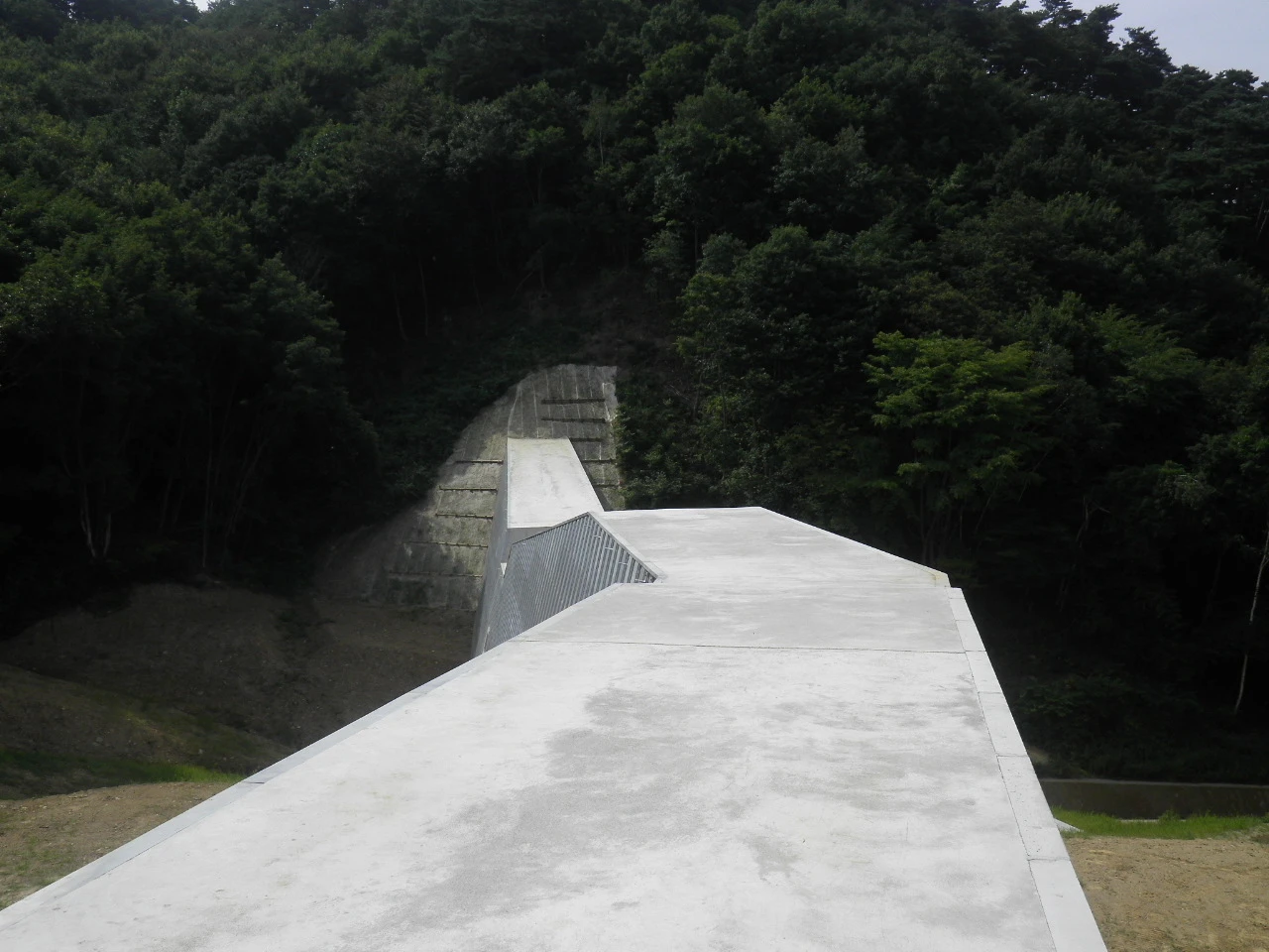 砂防堰堤の天端の上から見た、下流側へ下る袖部と山の斜面
