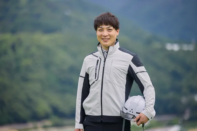 生坂村の山を背に、ヘルメットを持って立つ若手社員