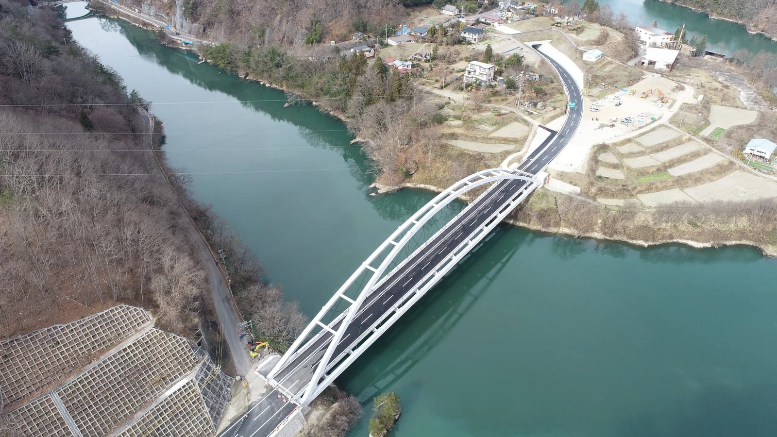 上空から見た、川に架かる白いアーチ橋と、対岸へ続く道路