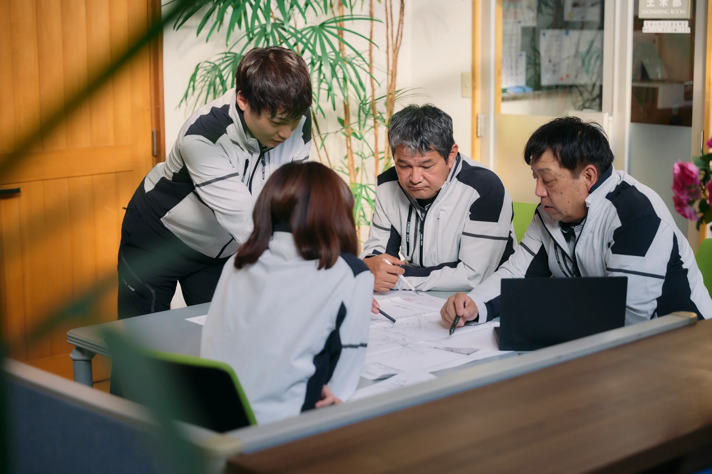 机を囲んで図面と資料を検討する4人の社員